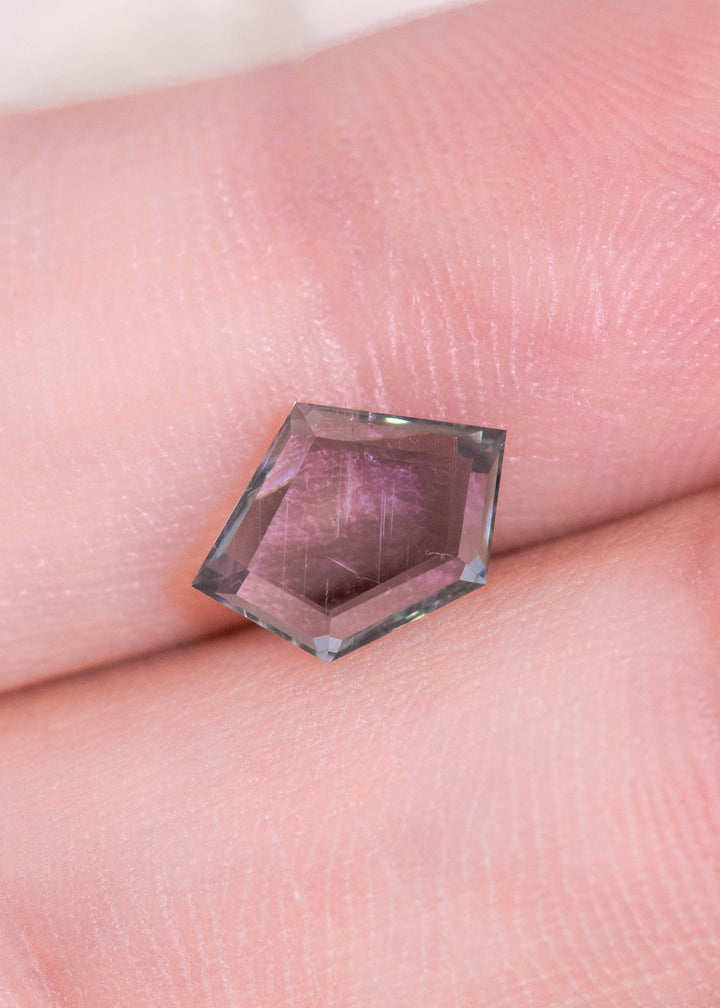 Macro fingertip scale photo of a natural Montana sapphire displaying purple, teal, and seafoam color shift, emphasizing the geometric pentagon portrait cut and precision faceting.