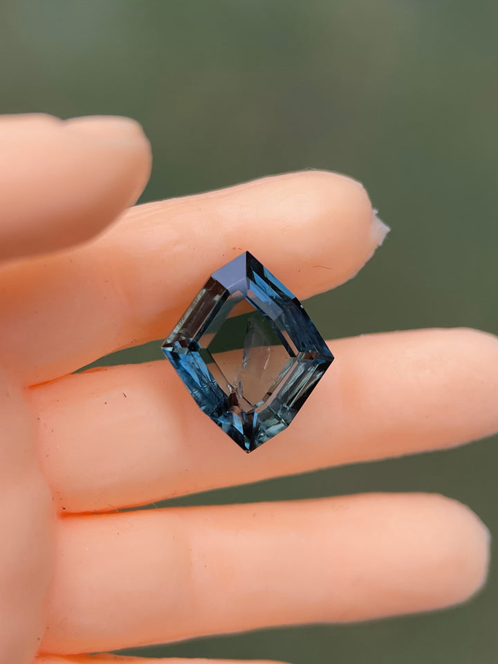 Detailed image of a 1.68-carat no heat sapphire, featuring a captivating teal and blue hue in a custom geometric rhombus portrait cut, held with precision to display its intricate angles and clarity.