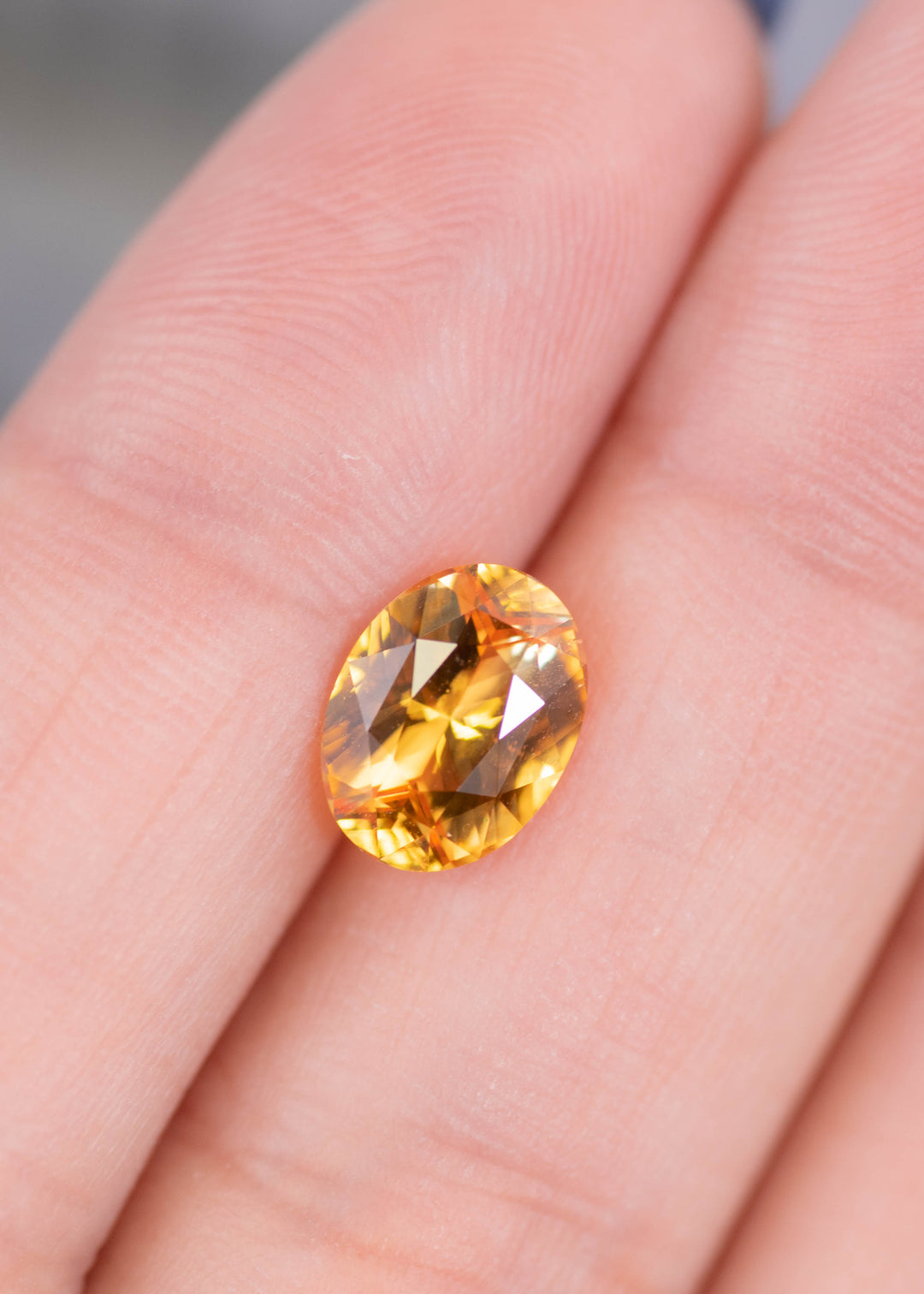 Oval cut orange red sapphire displayed on fingertips, showing scale, clarity, and warm golden color tones in a 2.53ct Sri Lankan sapphire