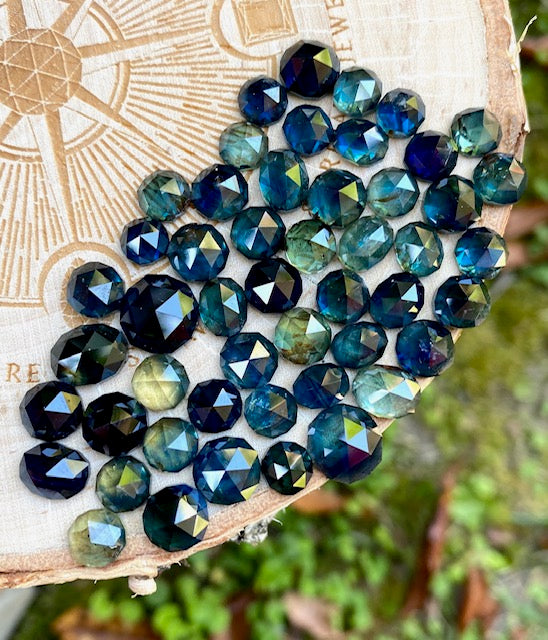 Blue, teal, and green rose cut loose sapphires arranged on wood, selected for custom jewelry accent stones and designer sourcing.