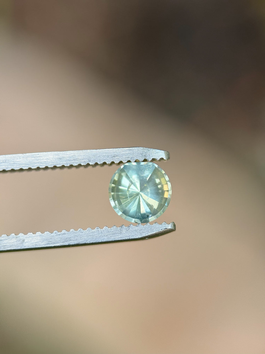 Close-up of a 0.96-carat unheated seafoam and yellow sapphire held in precision tweezers, showcasing its crisp moon phase round cut with a blurred brown-toned background.