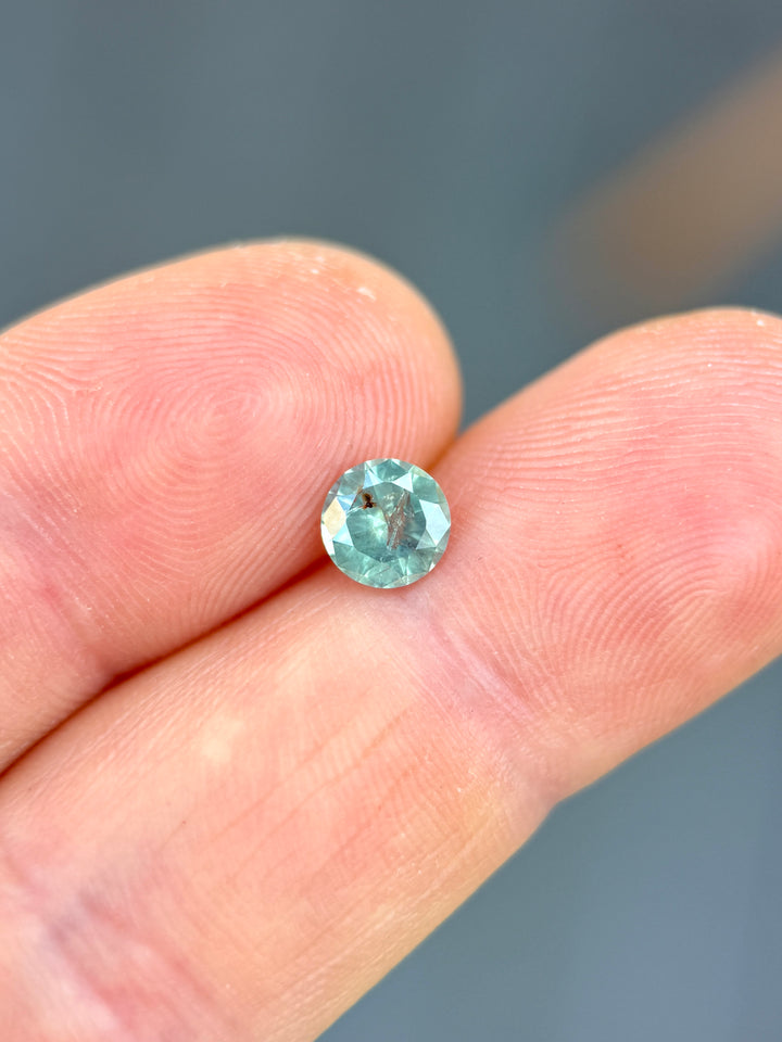 Round cut Montana sapphire resting on fingertips, showing opalescent powder teal tones and balanced brilliance.