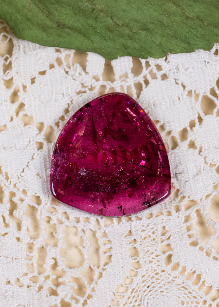 Close-up photograph of an untreated 28.02 carat Congolese pink tourmaline slice resting on cream lace beneath a green leaf. The polished gemstone displays saturated magenta and raspberry tones, natural dark inclusions, and a broad, softly triangular outline.