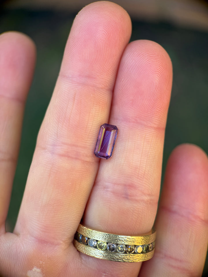 Natural 2.01 carat rutilated purple sapphire resting on fingertips for scale, precision custom elongated emerald portrait cut from Sakaraha, Madagascar, showing visible rutile inclusions and untreated crystal structure