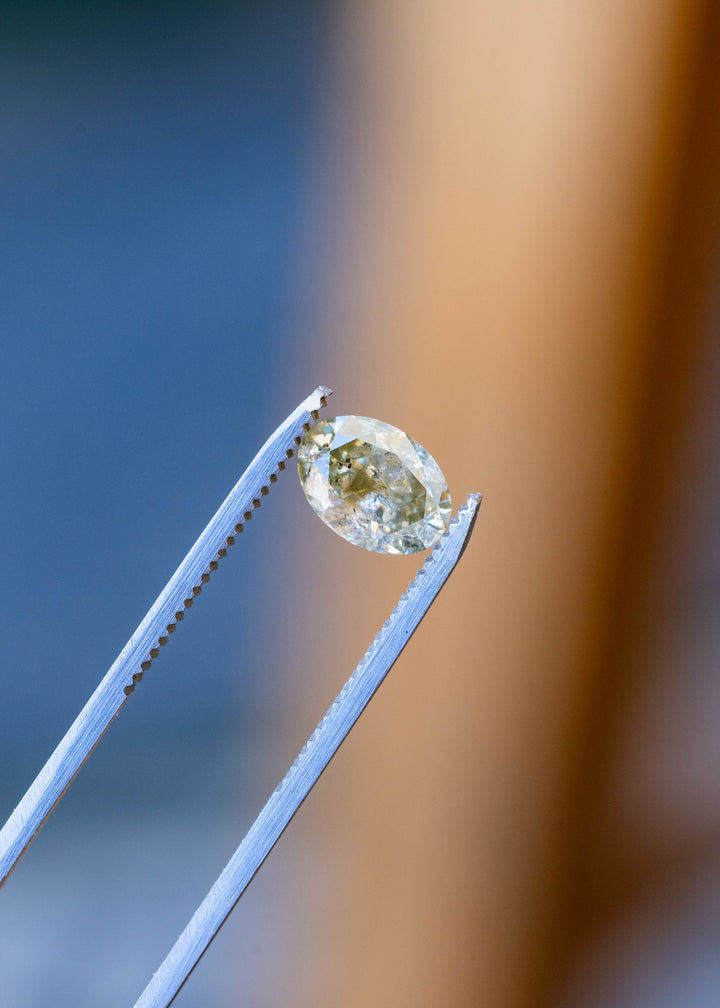 Fancy greenish brown grey salt and pepper oval diamond held in metal tweezers against a pale background, showing its brilliant faceting, elongated proportions and included interior.
