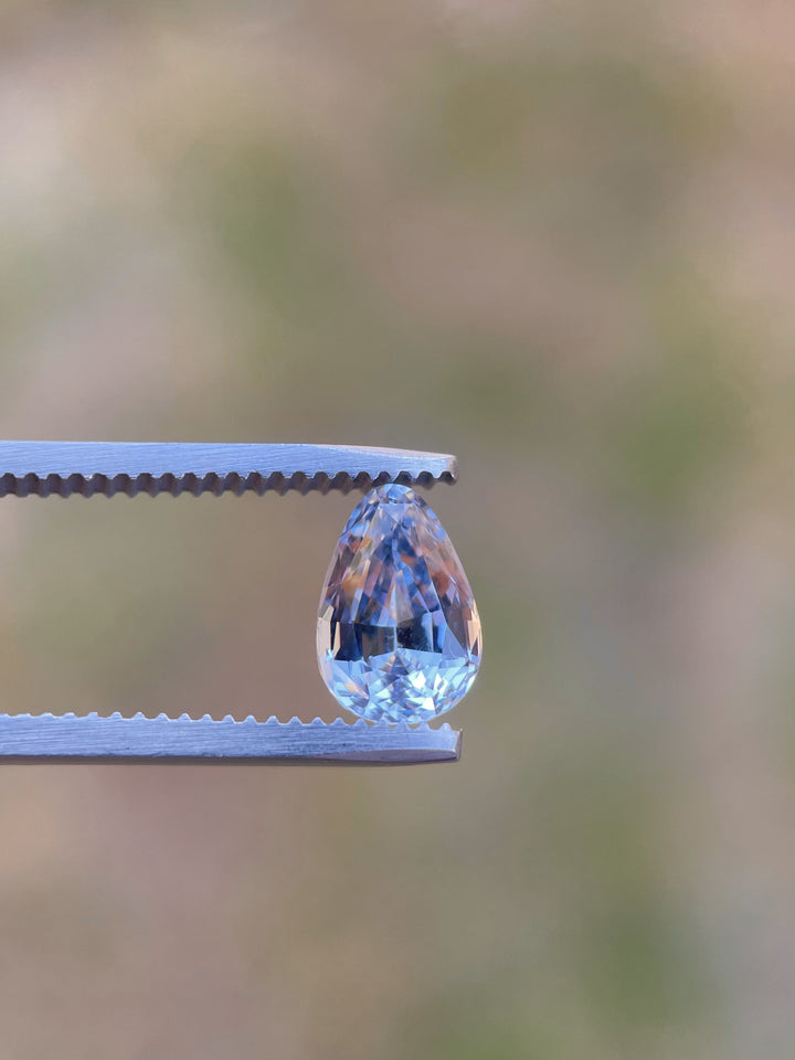 A finely cut 2.39-carat sapphire displayed in tweezers, with a mix of cornflower and powder blue shades, custom shaped into a brilliant teardrop. This untreated gemstone hails from Sri Lanka and glows against a subtle gray backdrop.