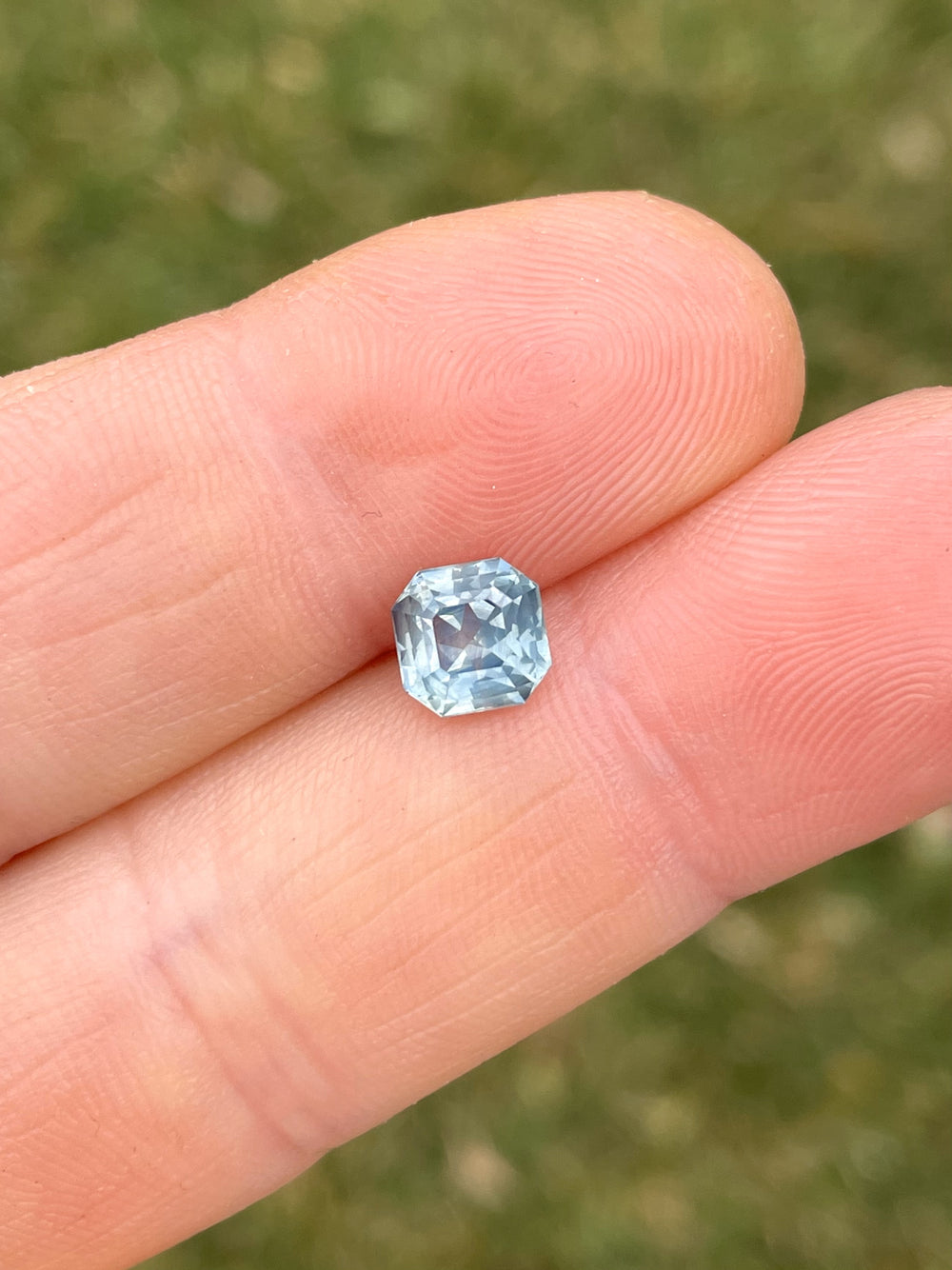 A beautiful 1.59-carat opalescent powder teal sapphire, octagonal step cut, displayed in a hand against a grassy background.
