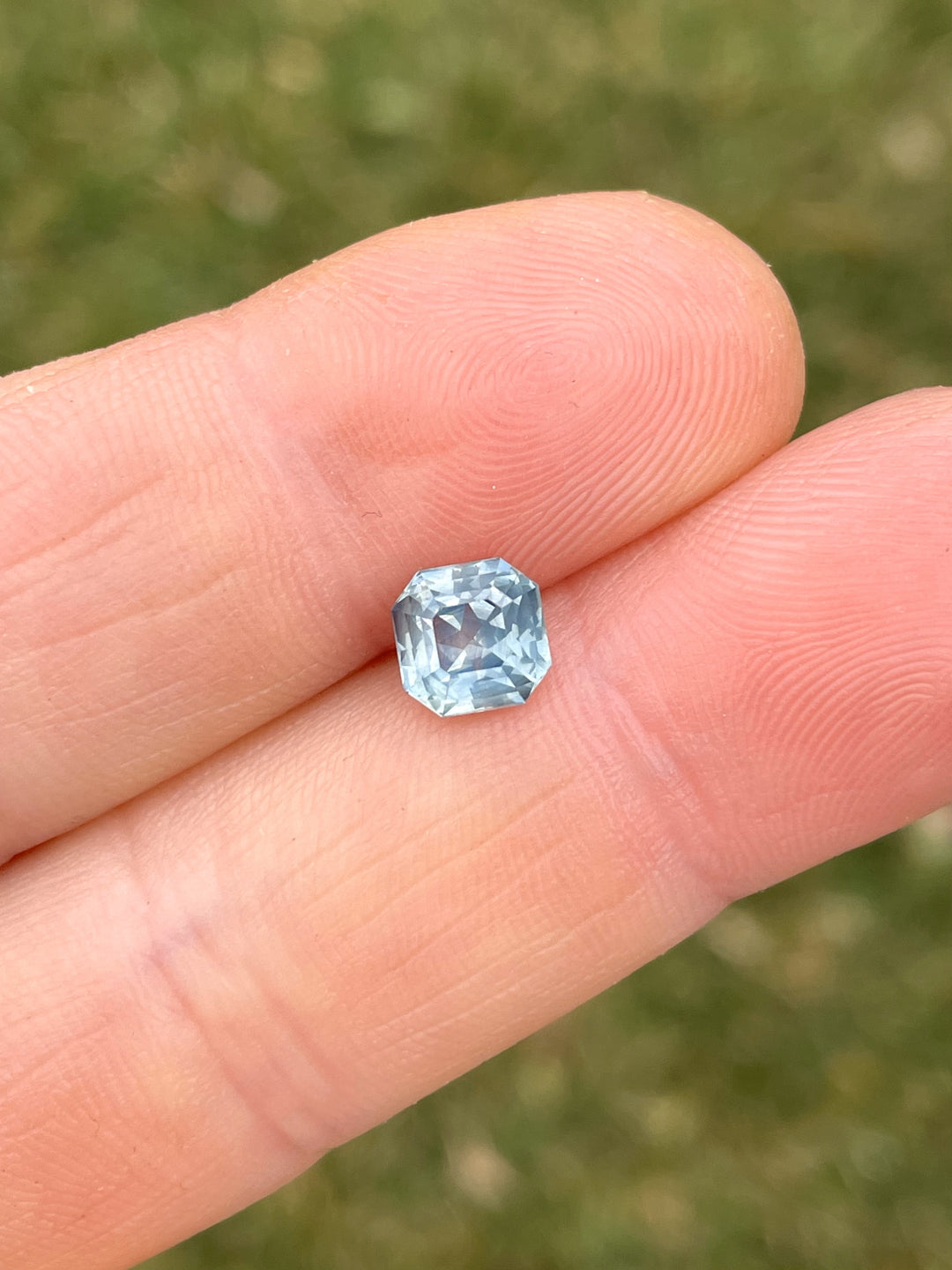 A beautiful 1.59-carat opalescent powder teal sapphire, octagonal step cut, displayed in a hand against a grassy background.