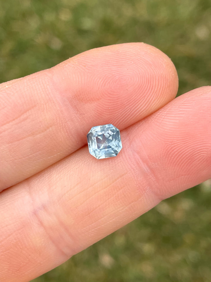 A beautiful 1.59-carat opalescent powder teal sapphire, octagonal step cut, displayed in a hand against a grassy background.