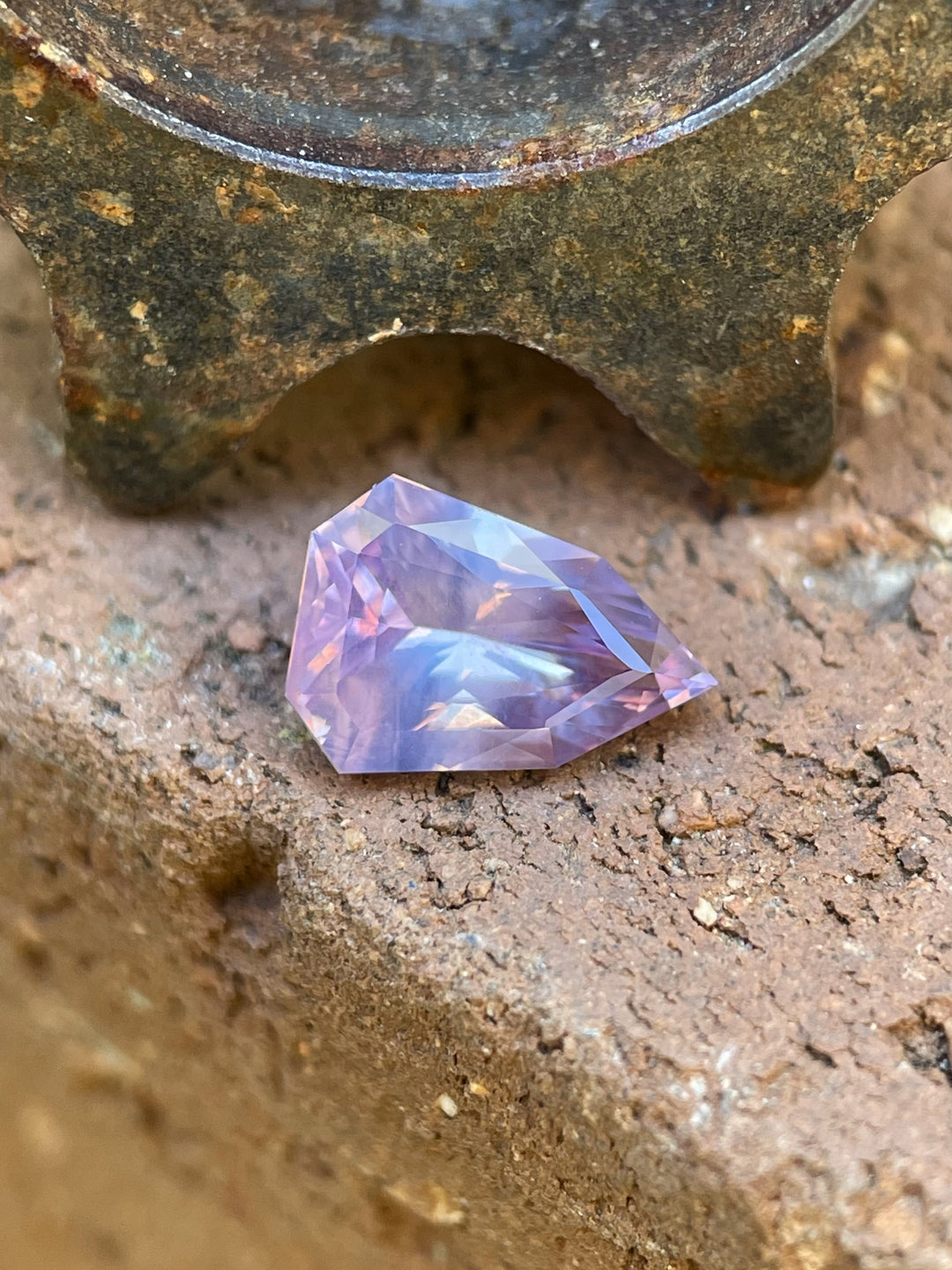 A close-up of a 1.54-carat pink and purple opalescent sapphire in a geometric shield cut resting on a weathered surface, showcasing its brilliance.