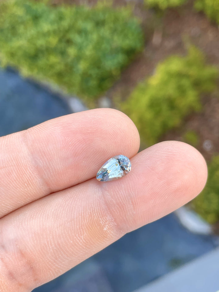 Close-up view of a hand holding a 2.00-carat mix custom teardrop sapphire, vividly displaying its seafoam and lime green hues. This Sri Lankan gem measures 10.30mm by 6.05mm by 4.44mm and is untreated, highlighting its natural beauty against a lush green background.