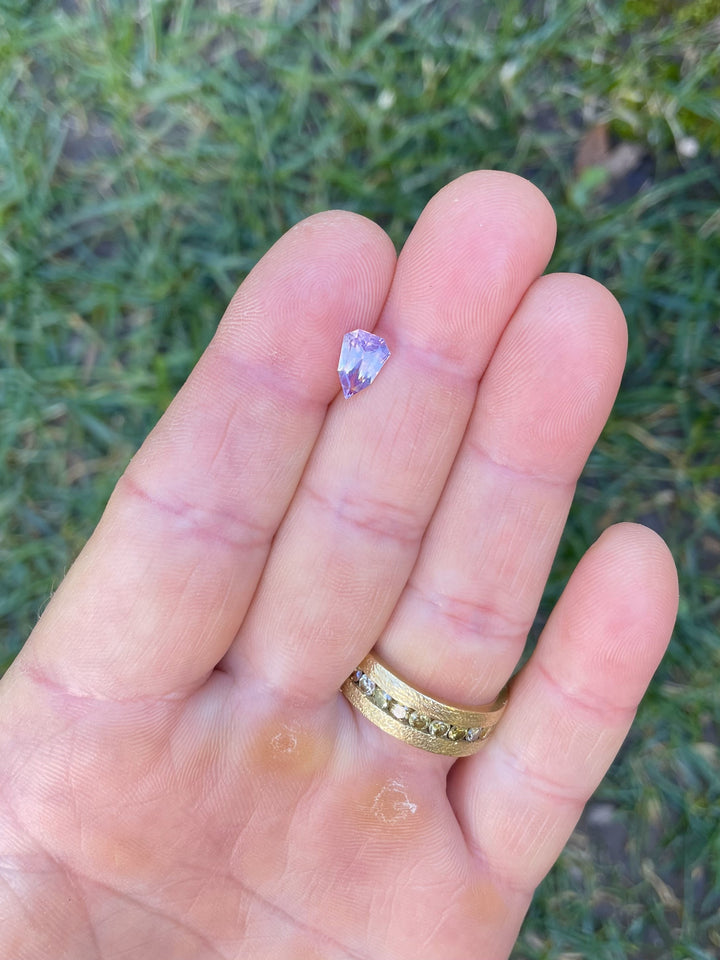 A 1.54-carat pink and purple opalescent sapphire in a custom geometric shield cut, displayed on a person's fingers with a grass background.