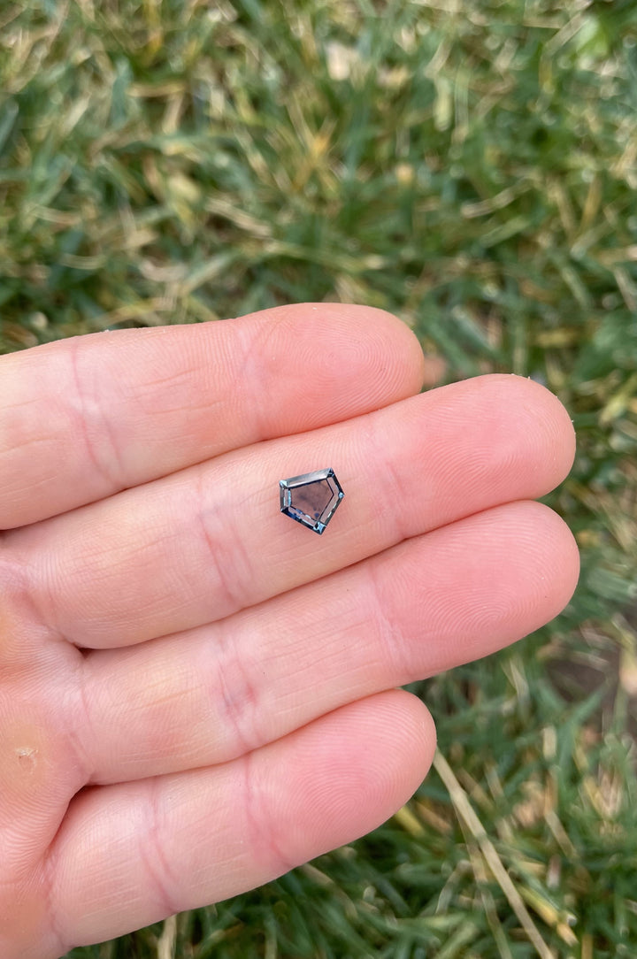 A custom step cut shield sapphire weighing 1.70 carats, resting on a fingertip, showcasing its parti-colors of blue, green, and yellow under natural lighting.