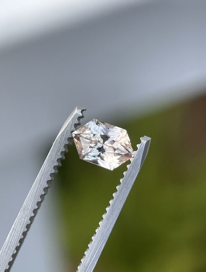 Peach champagne sapphire in hexagonal mix brilliant cut held with tweezers against a green blurred background.
