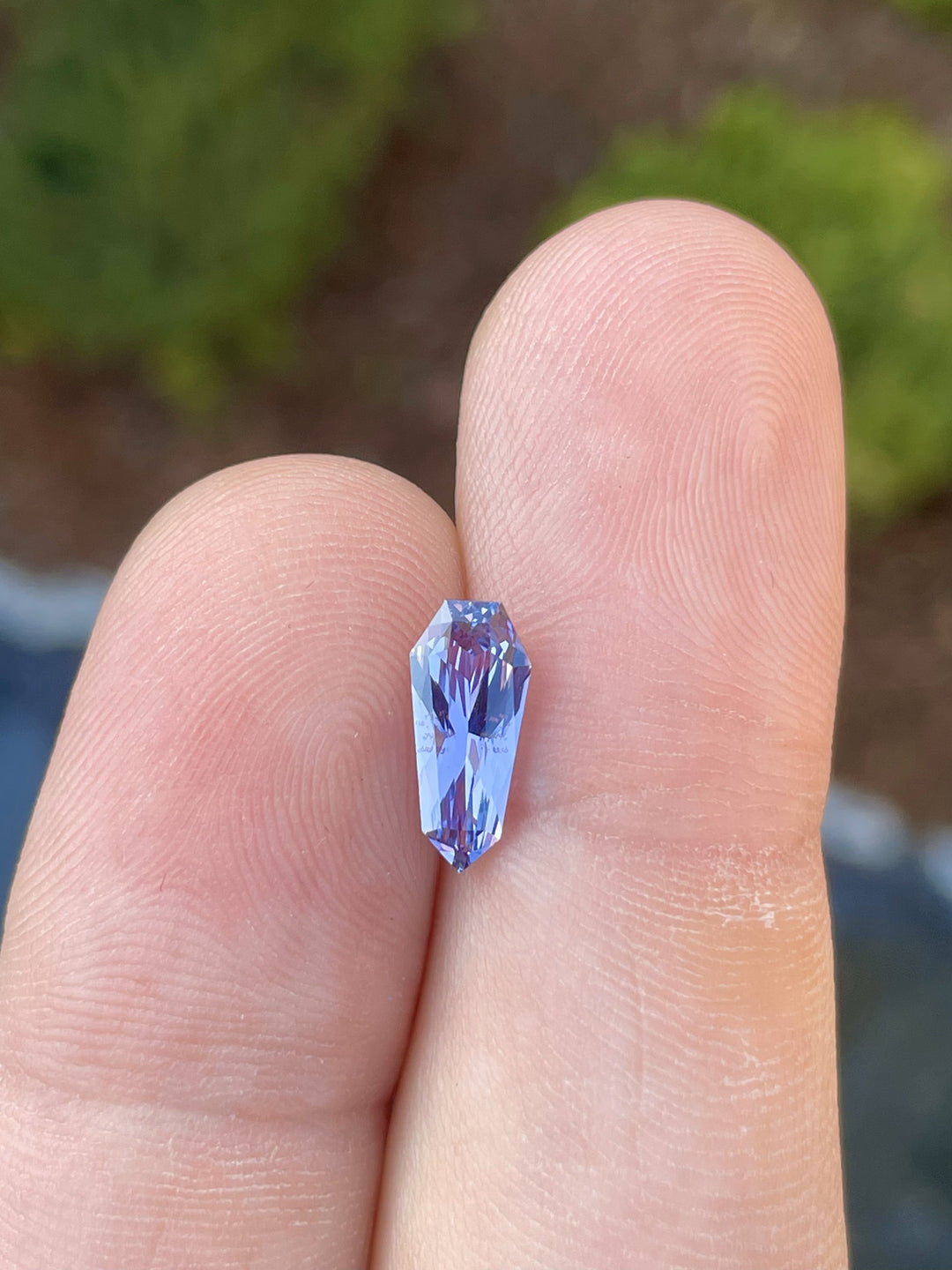 Close-up view of a 2.03-carat lavender, violet, and gray sapphire held between fingers, showcasing its intricate custom geometric shield cut and natural brilliance against a green background.