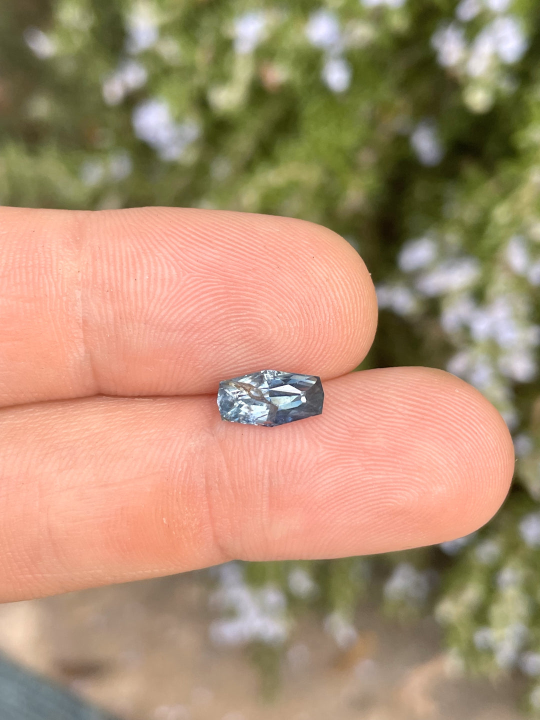 A 2.14-carat denim blue sapphire rests on a fingertip, showcasing its elongated cushion cut and deep blue hues. The untreated stone, sourced from the Umba River, Tanzania, measures 10.66mm by 5.55mm by 4.17mm and is captured against a blurred floral background.