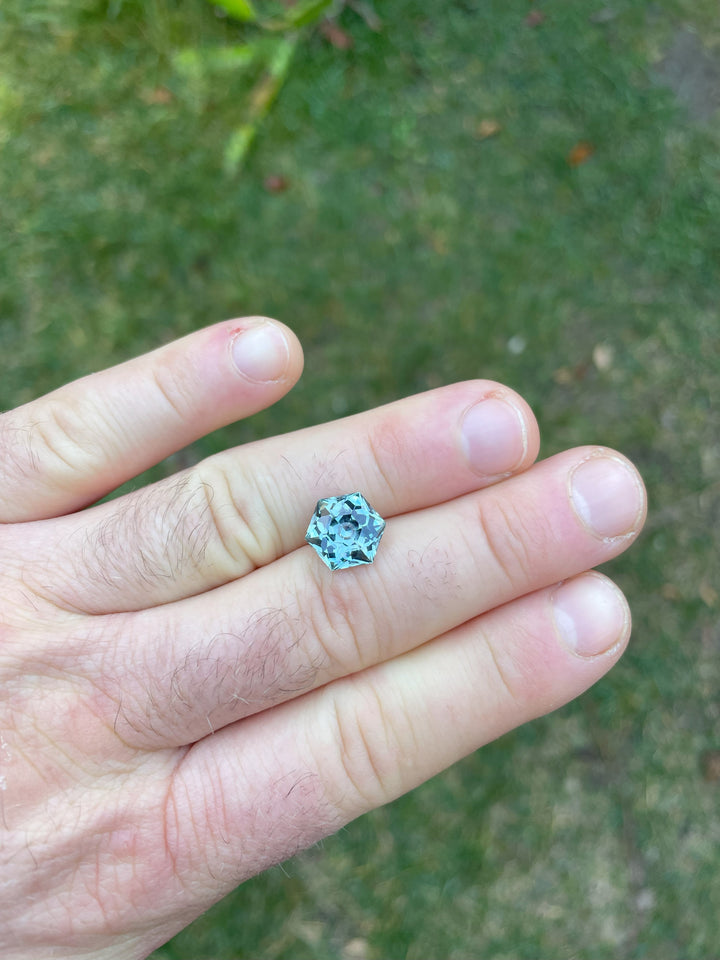 A striking 5.79-carat hexagonal modified brilliant sapphire resting on a finger, illuminated by natural daylight which enhances its green and blue hues, sourced from Montana, USA.