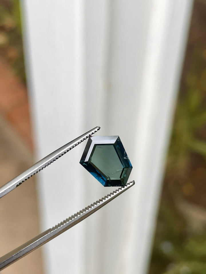 A 3.77-carat custom shield portrait cut sapphire held by tweezers, highlighting its deep peacock, teal, blue, and green colors. The stone, sourced from Garba Tula, Kenya, is untreated and measures 12.54mm by 9.29mm by 3.02mm, reflecting a vivid brilliance under natural light.