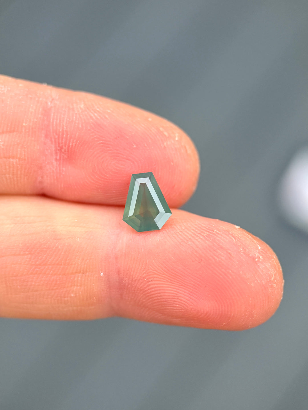 Shield step cut Kenyan sapphire resting on fingertips, showing layered teal, blue, and green tones with a soft opalescent glow.