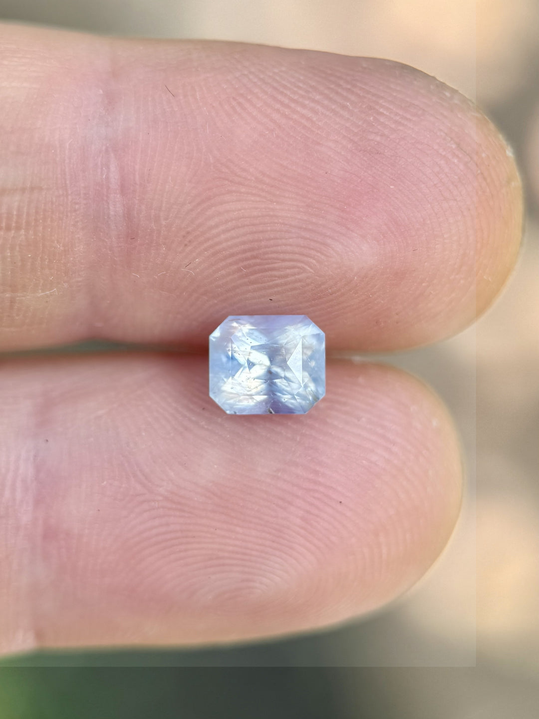 Radiant cut 1.58CT sapphire resting on fingertip, highlighting its translucent silver white and faint blue body
