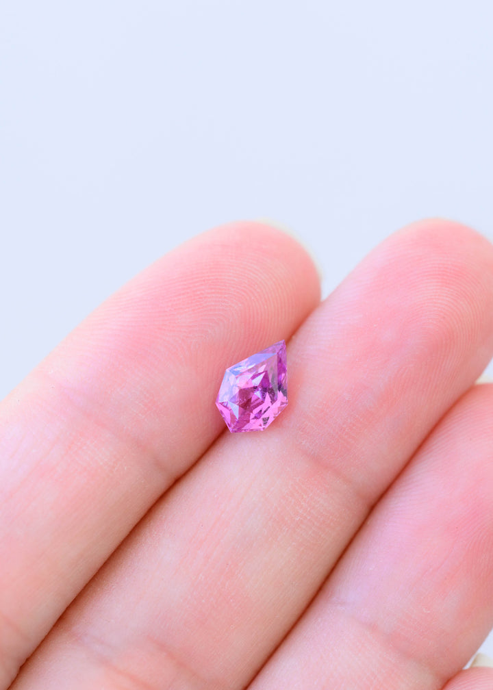 Small vivid pink and purple geo pear cut sapphire resting on fingertips for scale, showing its asymmetrical angular outline and bright reflective facets