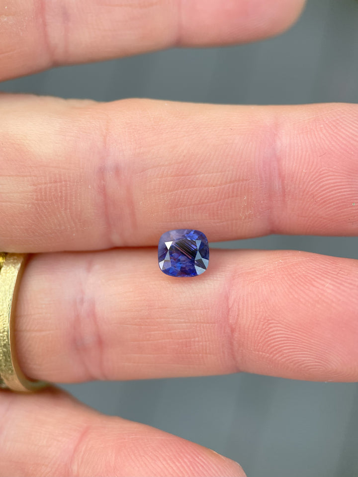 A 1.82-carat mix cushion cut sapphire featuring smokey lavender, violet, and parti colors, displayed on a person's finger.