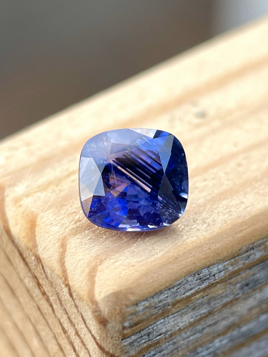 A mix cushion cut 1.82-carat sapphire in smokey lavender and violet colors, placed on a wooden surface with natural lighting.