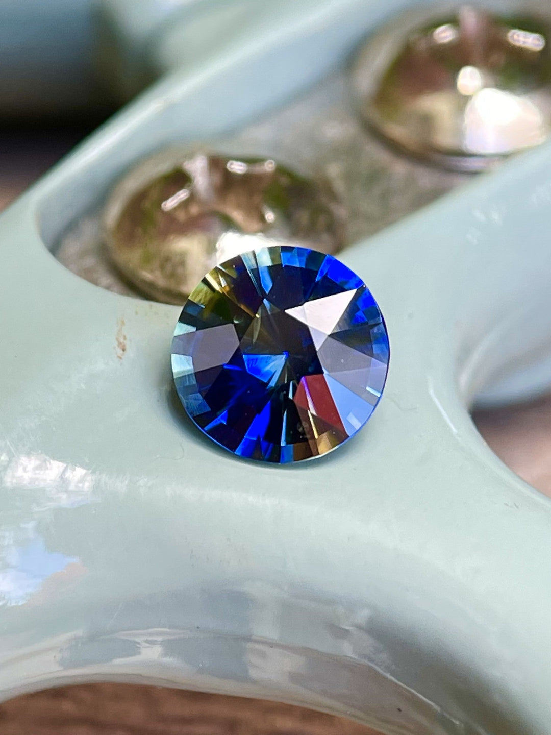 A 1.11-carat round brilliant parti sapphire with intense blue and yellow tones, resting on a glossy pale surface with metal accents in the background.