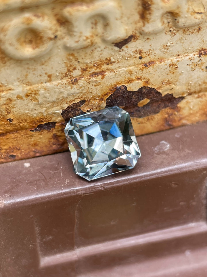 Close-up of a 1.58ct Montana sapphire in custom cut, showing its unique steely sage grey green tones and faceting against a rustic background.
