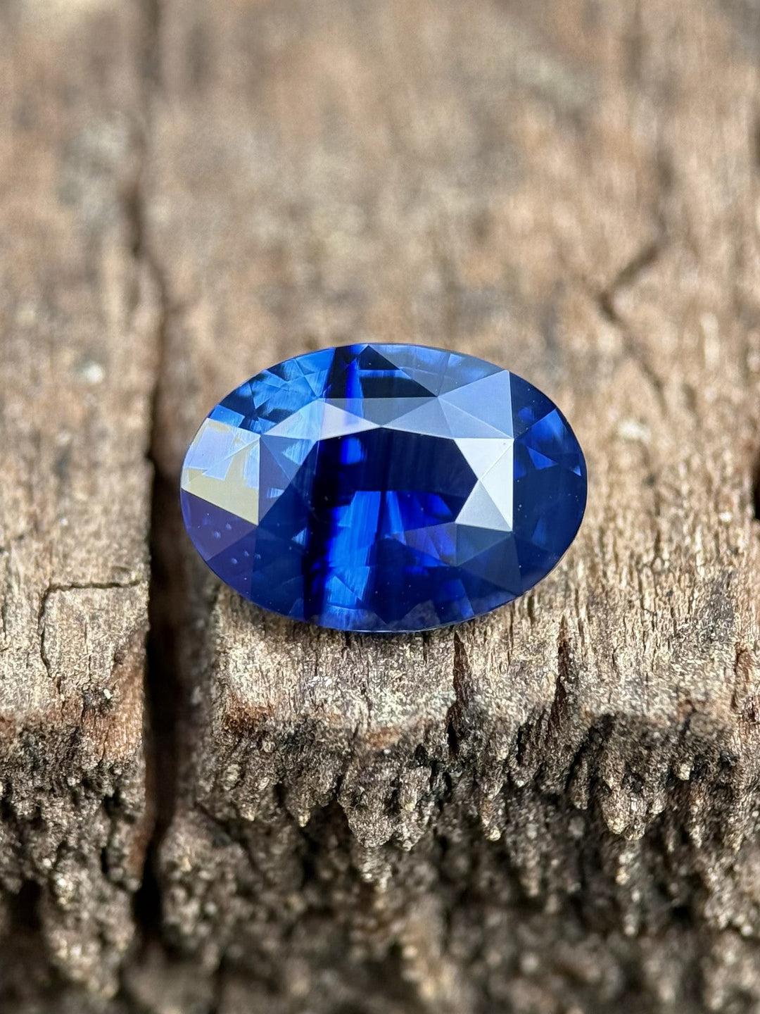 Striated royal blue parti sapphire, 2.56 carats, precision oval mixed cut, resting on textured wood surface while showcasing blue color zoning and brilliant facets from Diego, Madagascar, untreated.