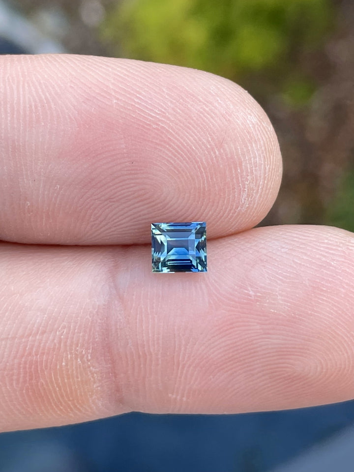 A rare 1.03-carat bi-color sapphire with green and blue hues, cut in an antique-style Carre shape, displayed on a fingertip.