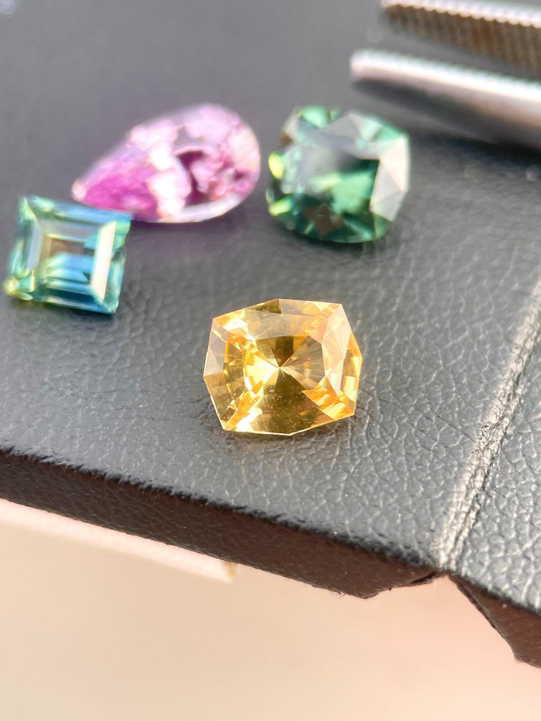 Close-up of a custom cushion cut sapphire with a vibrant blend of peach, apricot, and orange hues. This 1.52-carat gemstone, sourced from Sri Lanka, showcases a unique geometric shape with precise facets, resting on a textured black background.