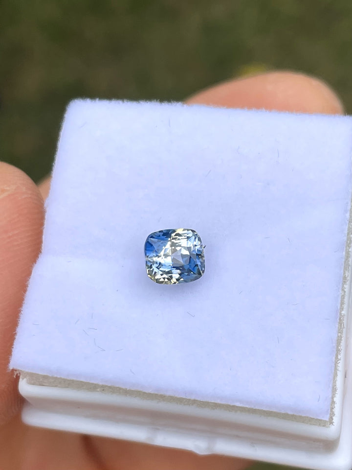 A 1.78-carat Madagascar Parti Sapphire carefully placed on a white display box. The modified brilliant cushion cut enhances its vibrant mix of yellow, colorless, denim, and Ceylon blue tones, reflecting light beautifully.