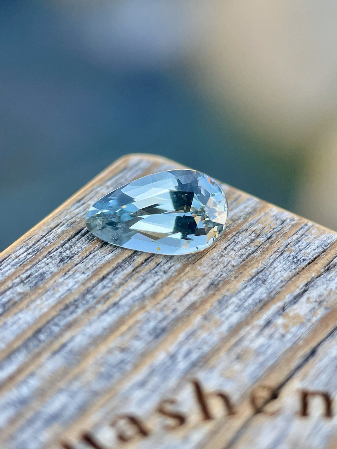 A 2.00-carat mix custom teardrop sapphire showcased on a wooden display stand with 'Quashen' engraved on it. The gemstone exhibits vibrant seafoam and lime green colors, measuring 10.30mm in length, 6.05mm in width, and 4.44mm in height, originating from Sri Lanka without any treatments.