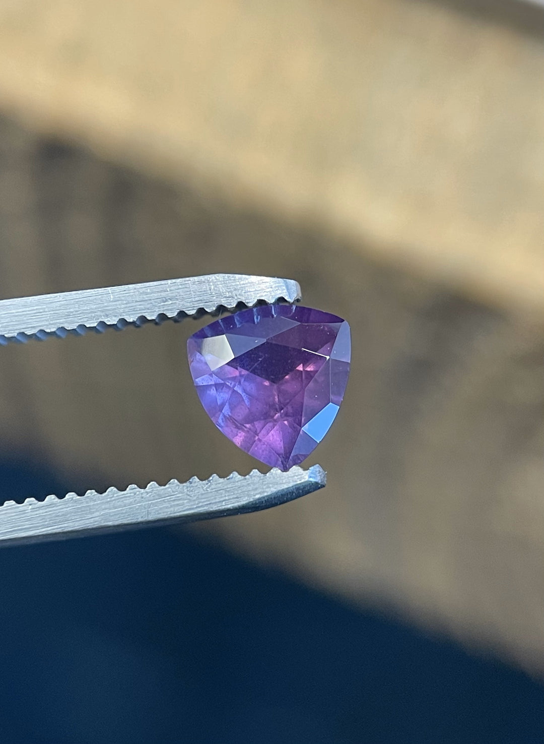 Vibrant 0.94ct trillion cut pink and purple sapphire from the Umba River, Tanzania, displayed in precision tweezers under directional lighting.