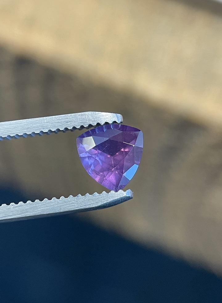 Vibrant 0.94ct trillion cut pink and purple sapphire from the Umba River, Tanzania, displayed in precision tweezers under directional lighting.
