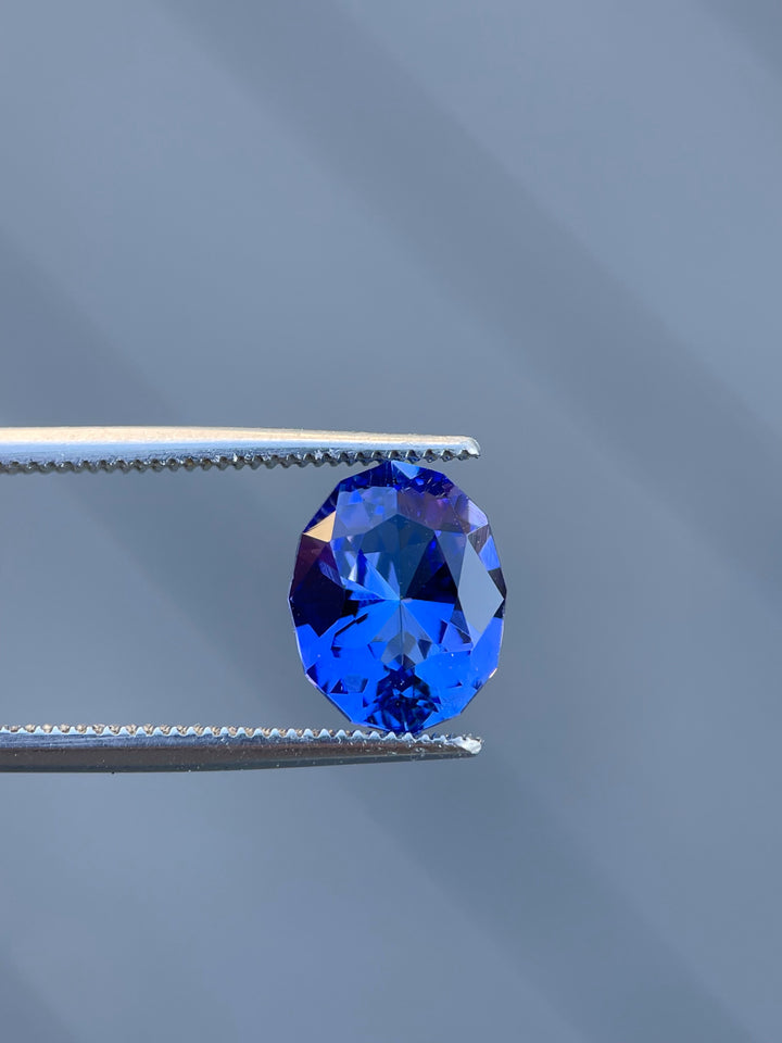 A dazzling 3.32 carat Tanzanite gemstone with a custom geometric oval cut, held by tweezers against a sleek background to highlight its violet and blue colors.