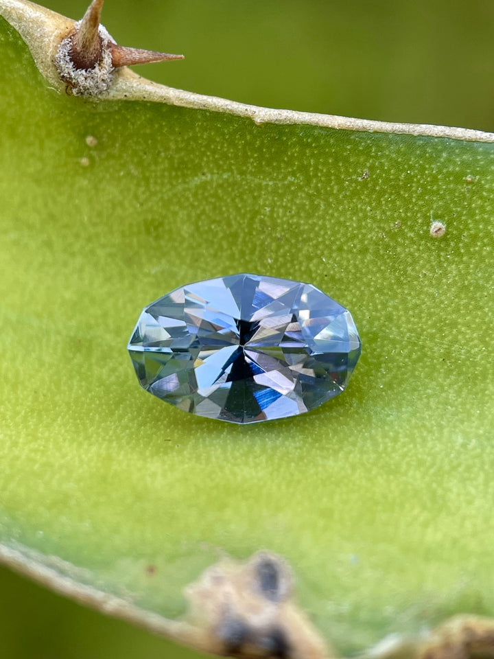 1.78ct Tanzanite with teal, lavender, and green tones resting on a natural leaf backdrop, precision-cut in a custom geometric oval shape.