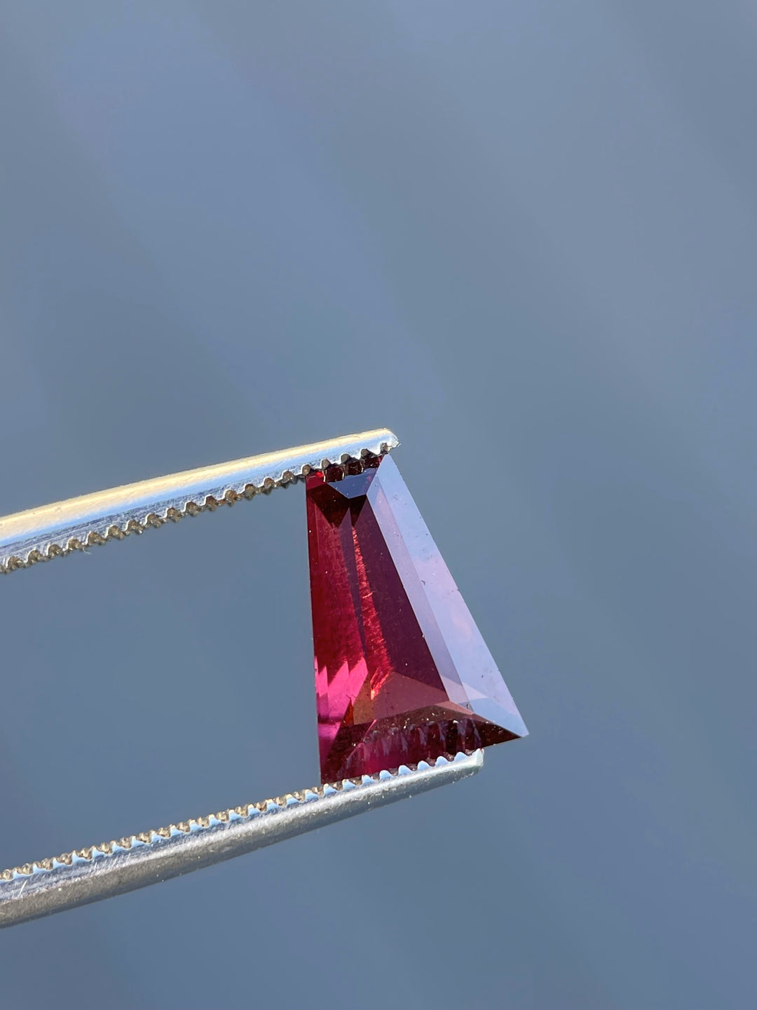 Garnet gemstone with tapered baguette cut, vivid red and purple tones, held with precision tweezers against a neutral background.
