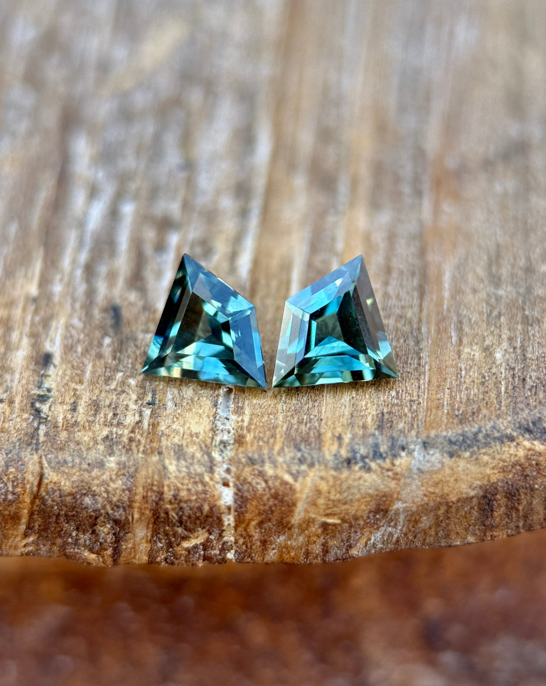 Teal and blue-green kite step cut sapphires standing upright on a textured wood surface, highlighting their geometric facets and natural unheated color from the Queensland Gemfields, Australia.