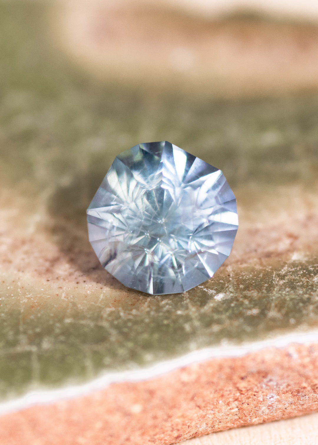 Teal blue green Montana sapphire resting on layered natural stone, showcasing a 1.40 carat untreated sapphire with custom modified round cut and crisp facet symmetry