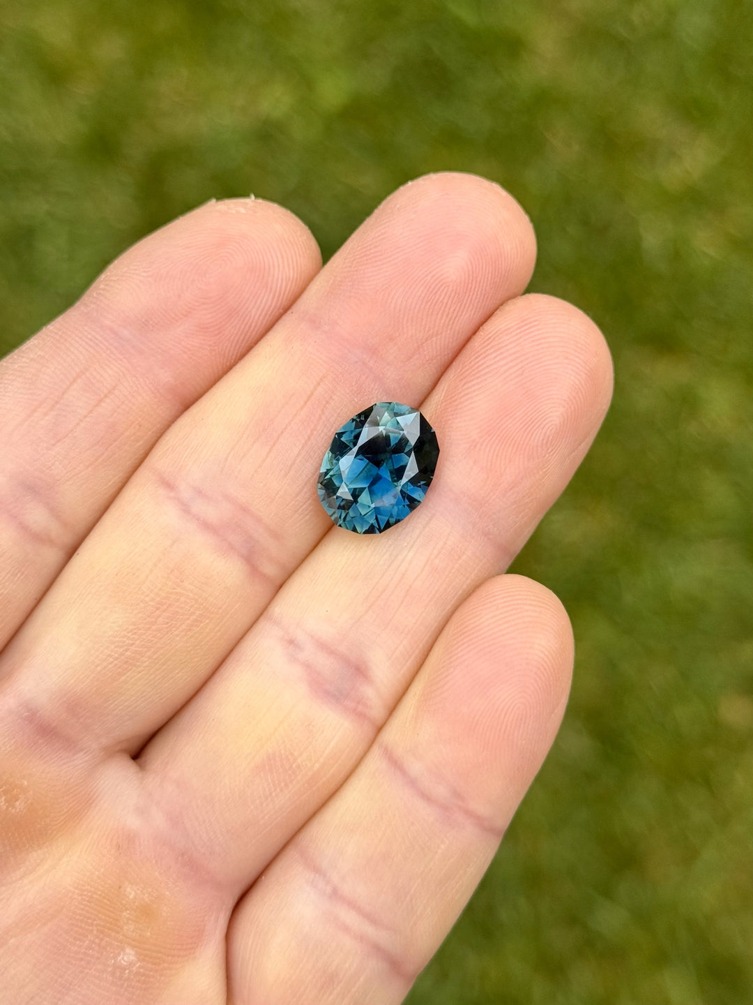 Close-up image of a 7.75-carat oval brilliant sapphire with teal and blue tones, shown resting on fingertips to highlight size and clarity.