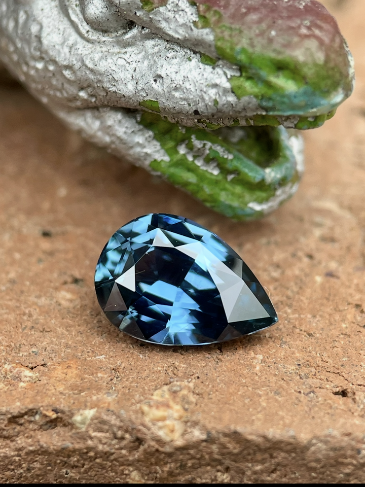 Close-up image of a 2.54 carat unheated sapphire from Madagascar, in a mix pear brilliant cut with teal, blue, and green hues, resting on a textured natural surface.