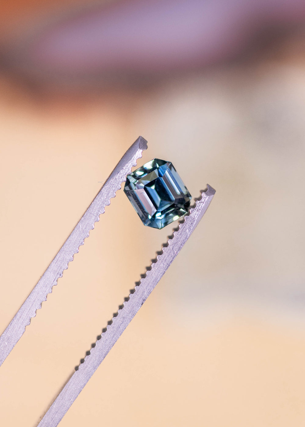Loose blue green emerald cut sapphire held in tweezers, showing crisp step facets and natural color zoning from Sakaraha, Madagascar.