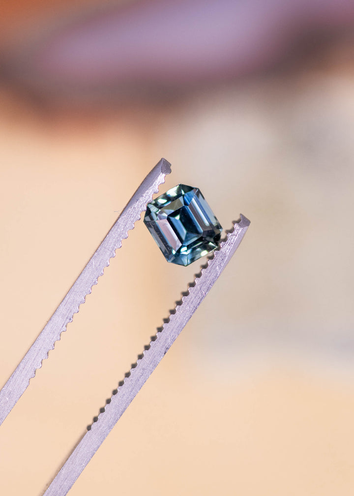 Loose blue green emerald cut sapphire held in tweezers, showing crisp step facets and natural color zoning from Sakaraha, Madagascar.