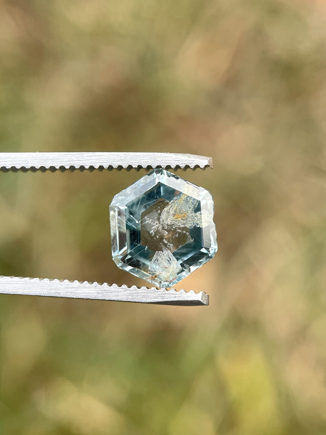 Natural teal green sapphire in hexagon portrait cut, captured with tweezers against a soft background.