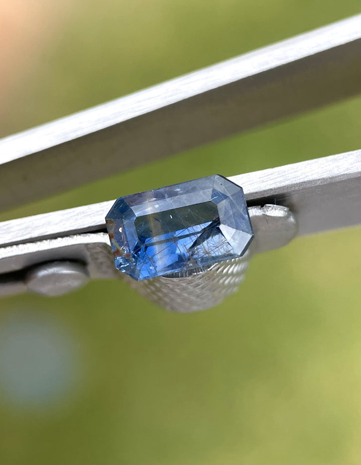 A close-up of a 1.45-carat sapphire with teal and blue parti hues, held in precision tweezers, showcasing its modified emerald cut and intricate details.