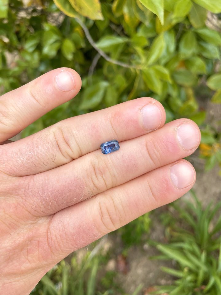 A 1.45-carat sapphire with teal and blue parti colors in a modified emerald cut, displayed on a finger with a lush green background.