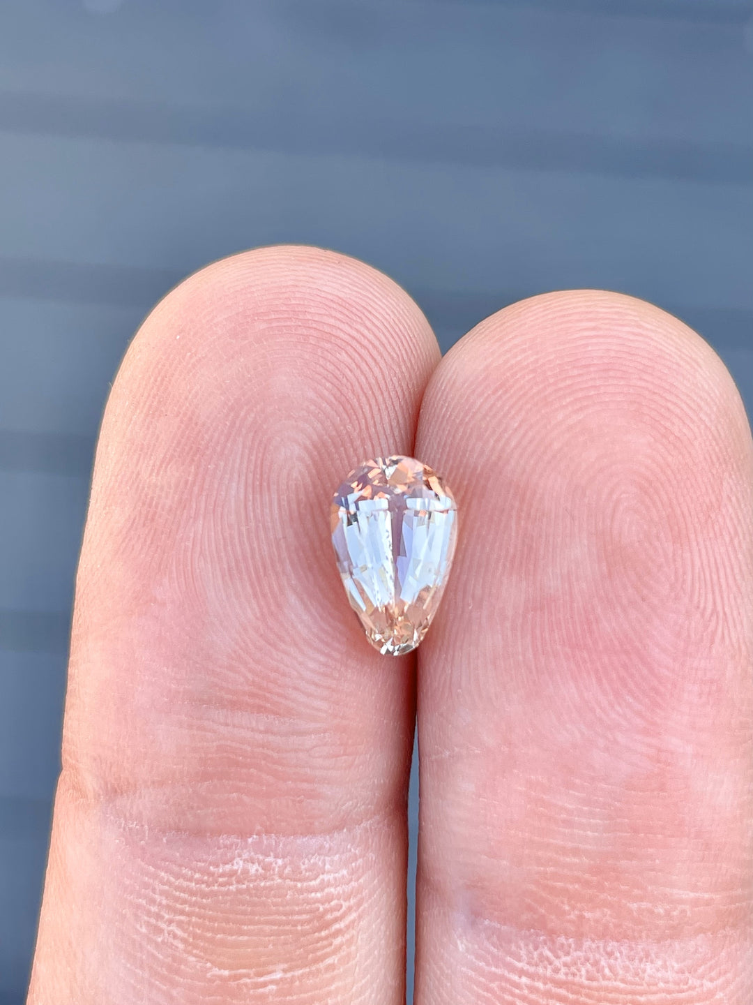 A 2.45-carat teardrop-shaped sapphire with pastel champagne and peach tones, shown resting on the fingertips of a hand for size comparison. The custom brilliant cut of the gemstone emphasizes its clean facets and luminous glow. Measuring 10.12mm by 6.40mm by 4.70mm, the sapphire originates from Sri Lanka and is untreated.
