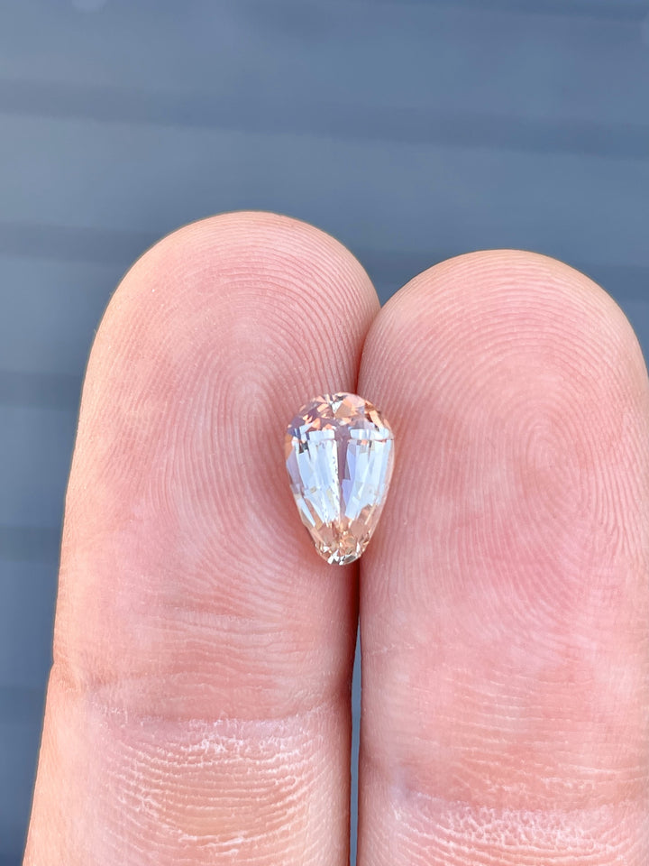 A 2.45-carat teardrop-shaped sapphire with pastel champagne and peach tones, shown resting on the fingertips of a hand for size comparison. The custom brilliant cut of the gemstone emphasizes its clean facets and luminous glow. Measuring 10.12mm by 6.40mm by 4.70mm, the sapphire originates from Sri Lanka and is untreated.